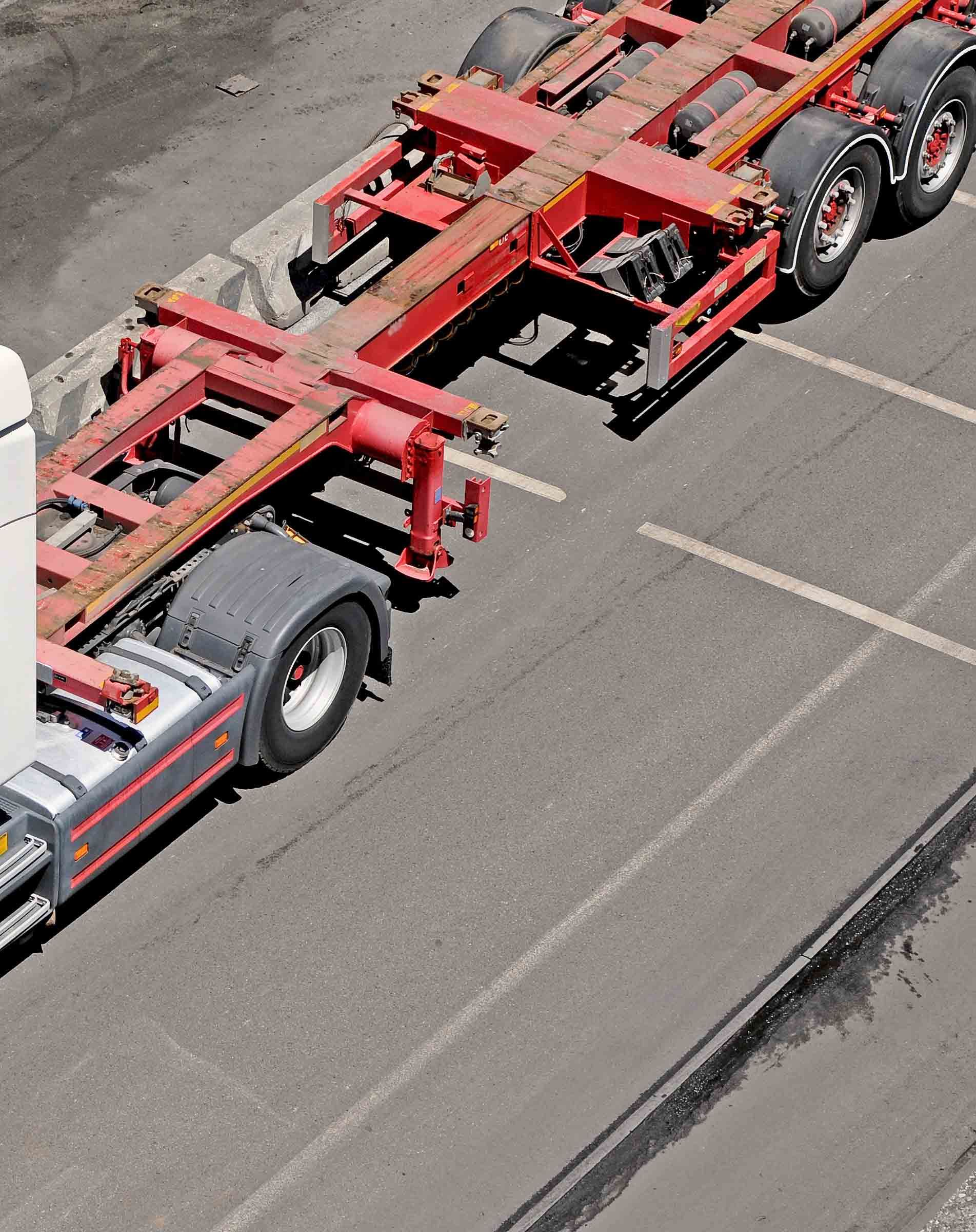 Red and silver truck hauling an empty container chassis on asphalt parking lot.