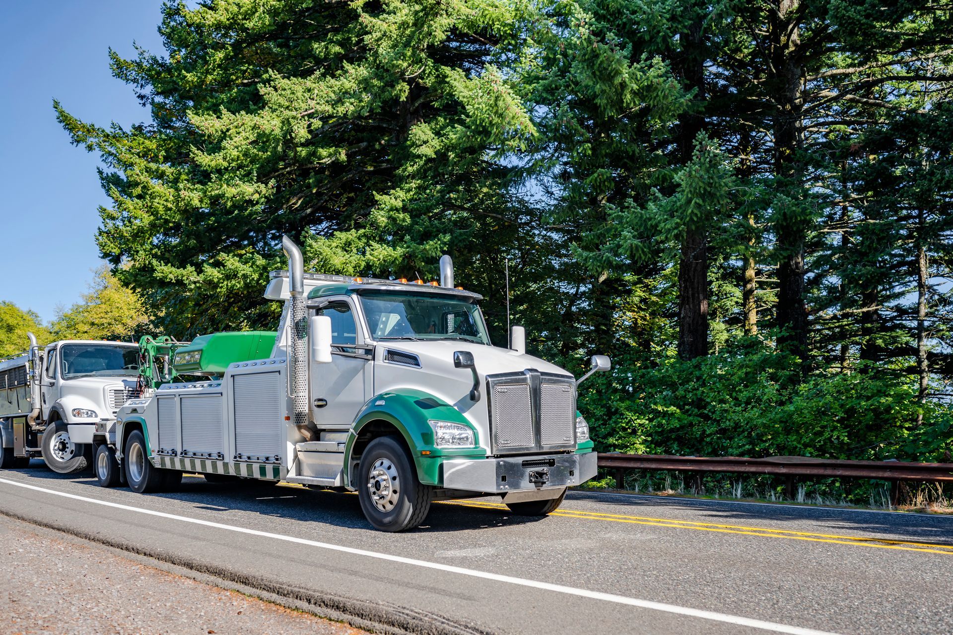 Tow truck towing a car on a road, trees in the background. Truck is white with green accents. Tow truck towing a car on a road, trees in the background. Truck is white with green accents.