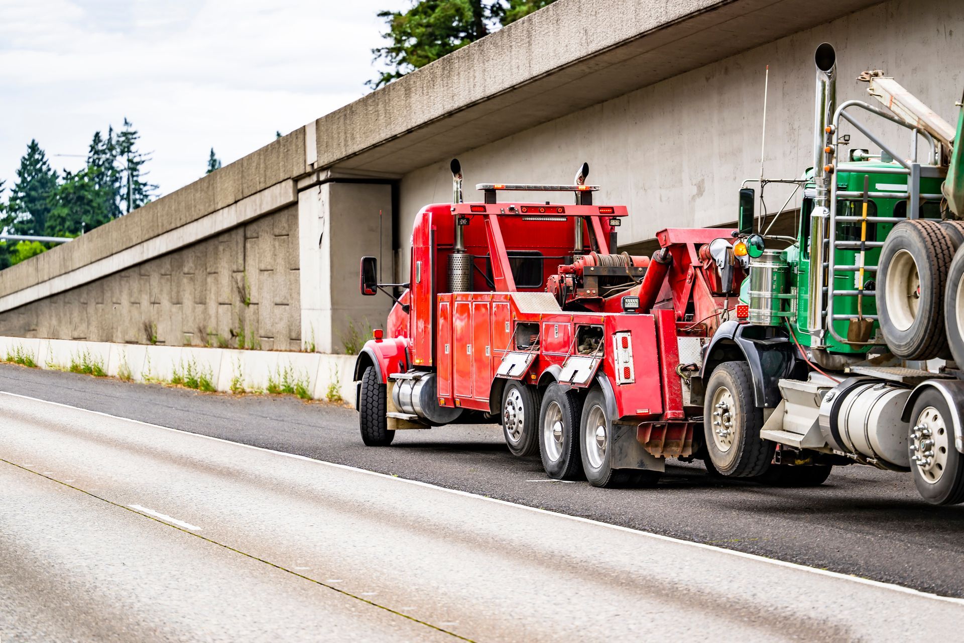 Red tow truck towing a green semi-truck on a highway, concrete barrier in the background. Red tow truck towing a green semi-truck on a highway, concrete barrier in the background.
