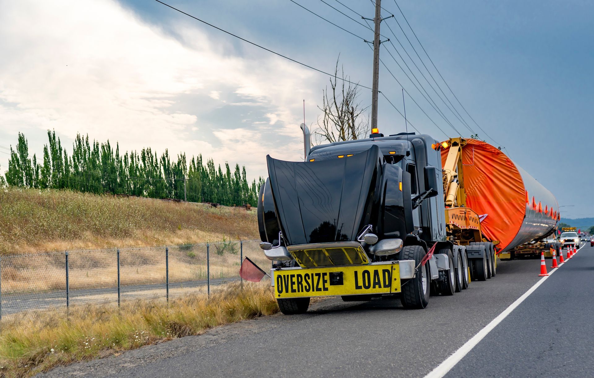 Oversize load truck on a road with a large orange cylindrical object. Black truck, yellow warning sign. Cloudy sky. Oversize load truck on a road with a large orange cylindrical object. Black truck, yellow warning sign. Cloudy sky.