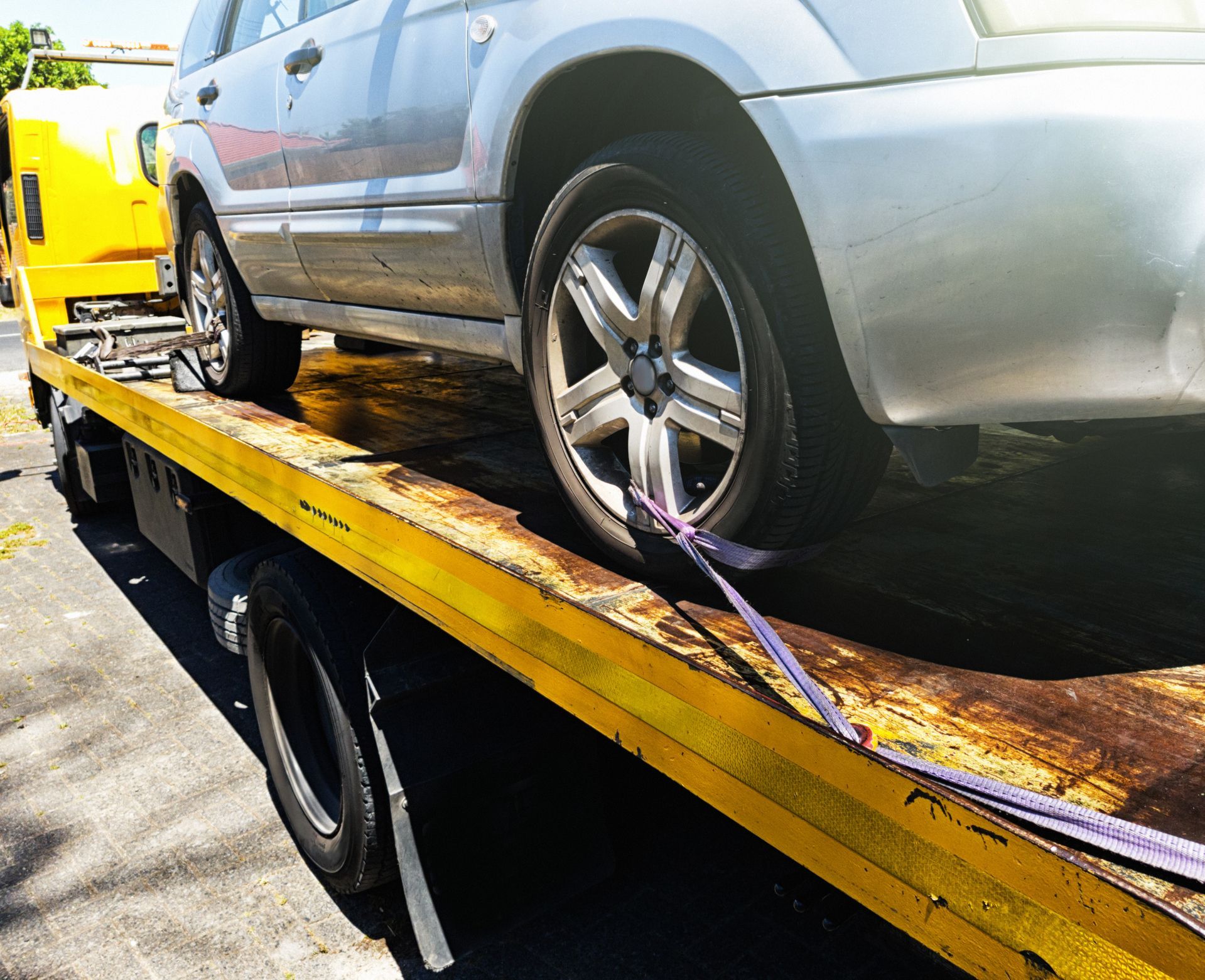 Silver car secured on a yellow tow truck bed with purple straps.