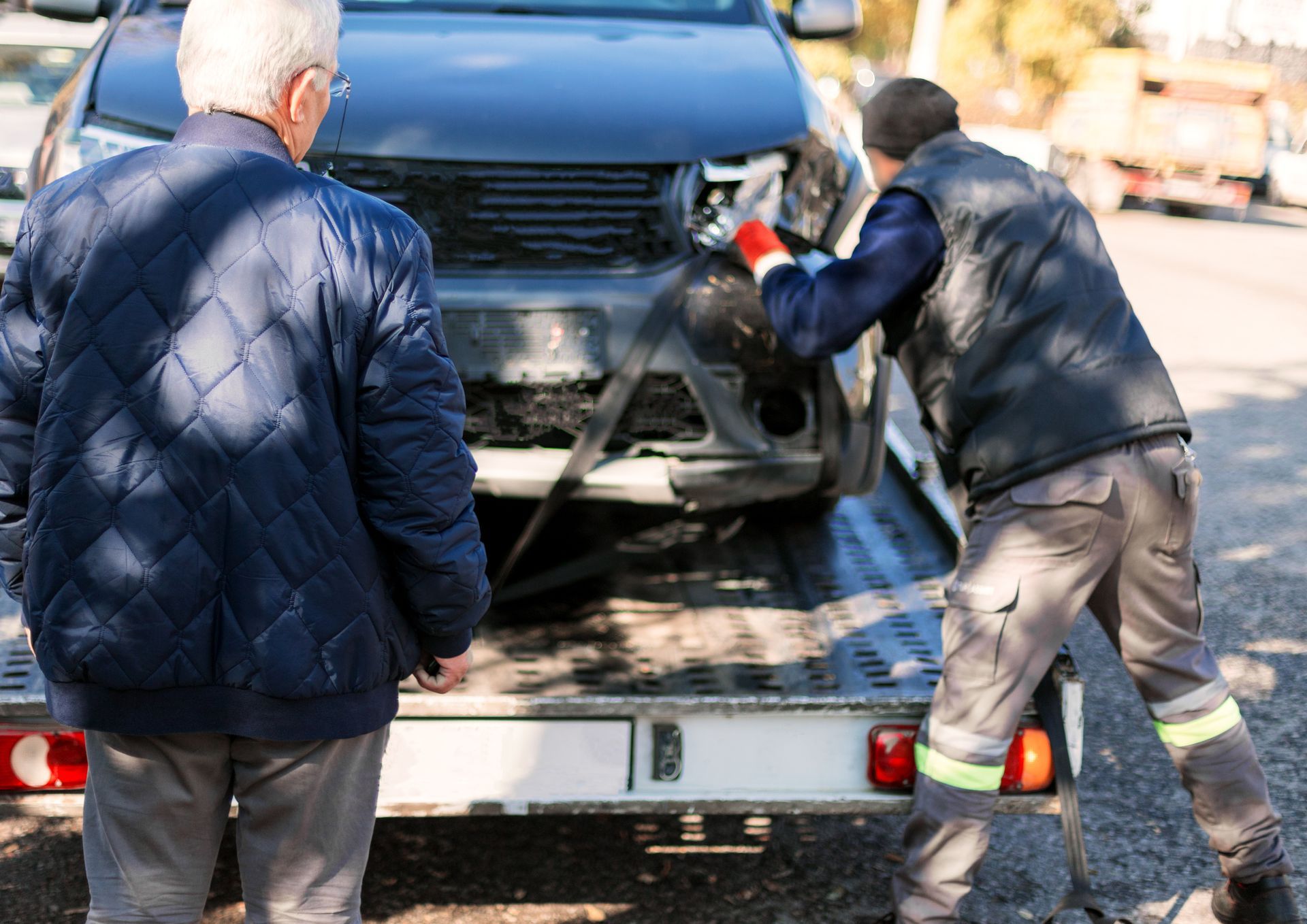 Man watches as a tow truck operator secures a damaged car on a flatbed trailer in a sunny street setting.