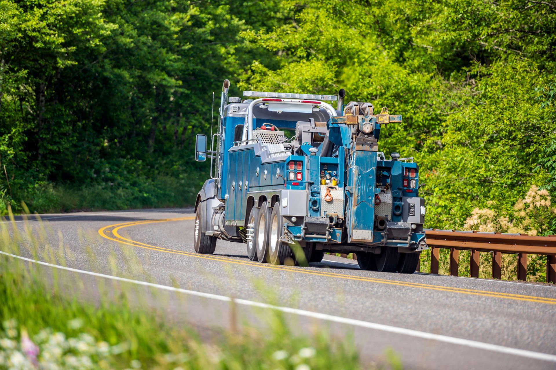 Blue tow truck driving on a curved road, surrounded by green trees and vegetation. Blue tow truck driving on a curved road, surrounded by green trees and vegetation.