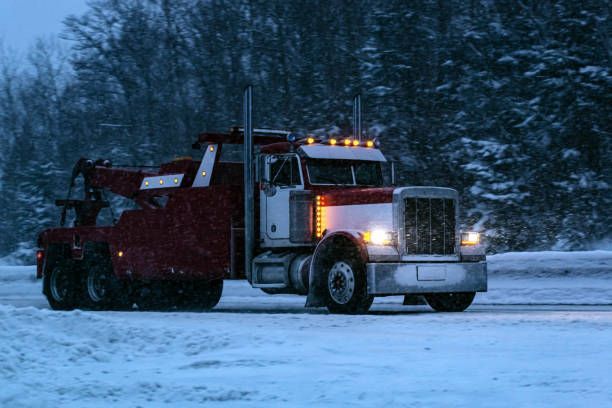 Tow truck on a snowy road, red and white colors, with headlights on, evening setting.