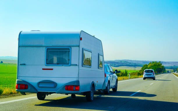 A car towing a white camper on a sunny road, with green fields on one side.