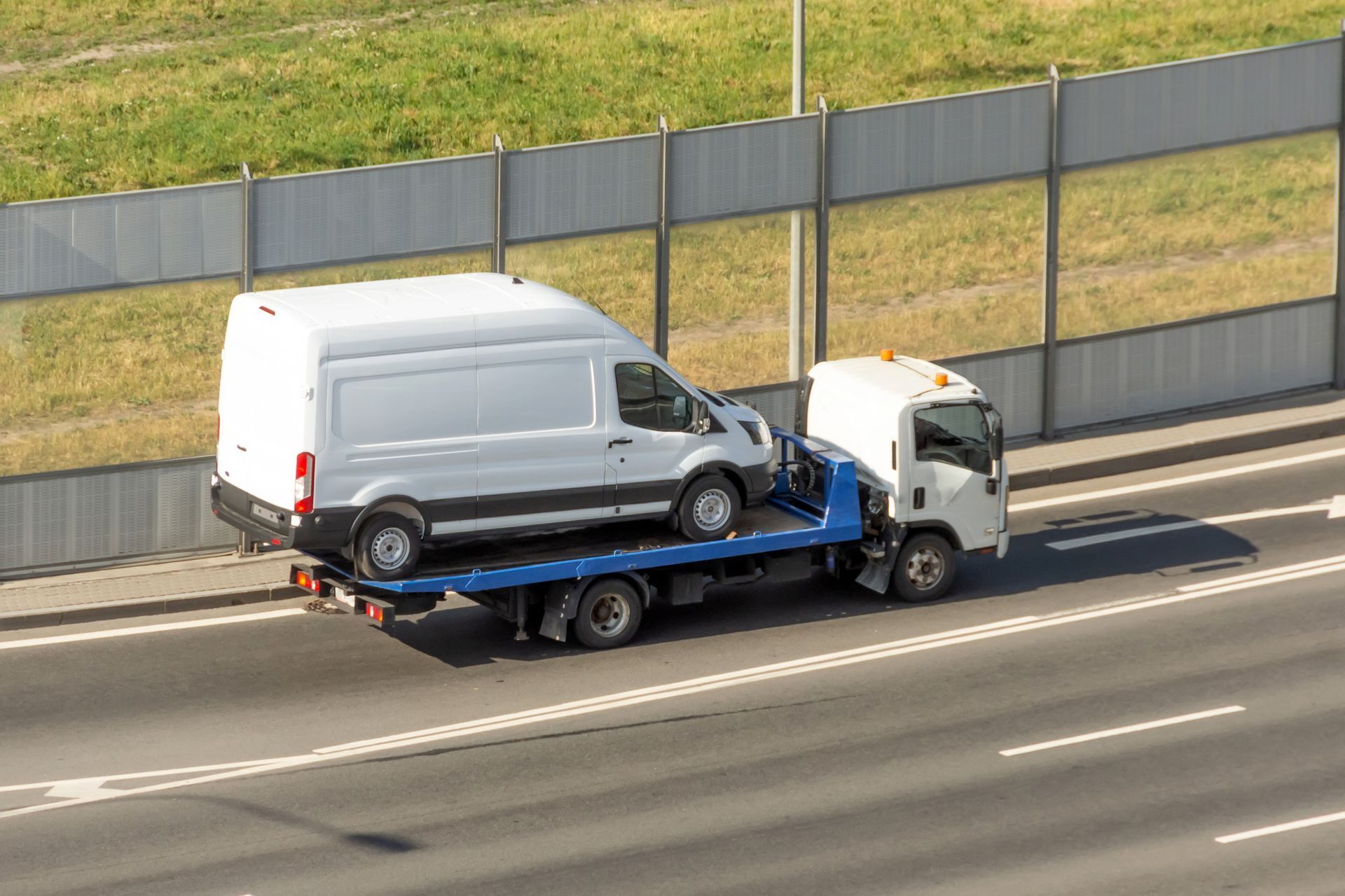 White van being towed on a flatbed truck on a highway, by a sound barrier.