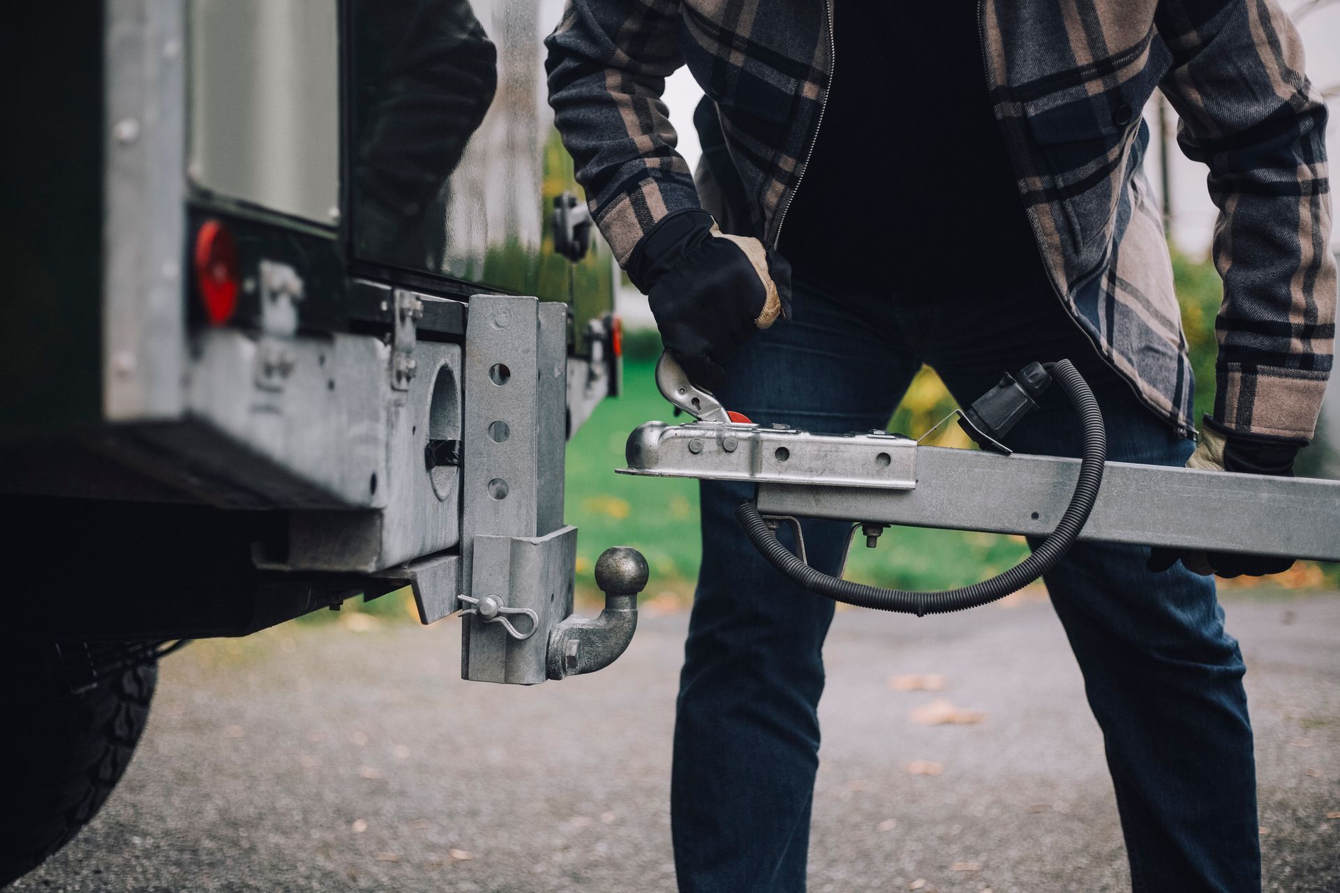 Person attaching a trailer to a vehicle hitch outdoors. Gray trailer and hitch, with a green lawn in the background.