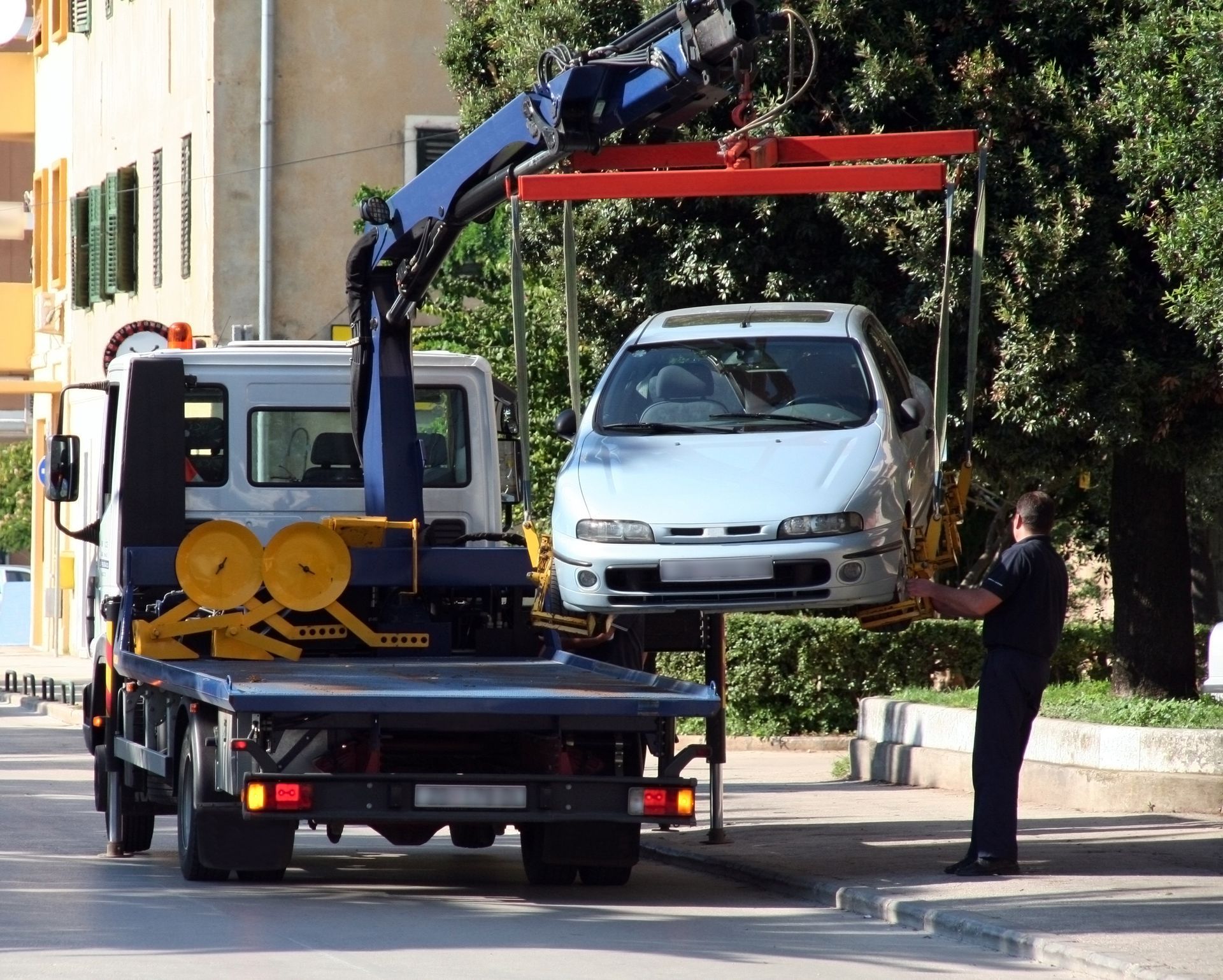 Tow truck lifting a silver car in a street. A worker stands nearby, overseeing the process.