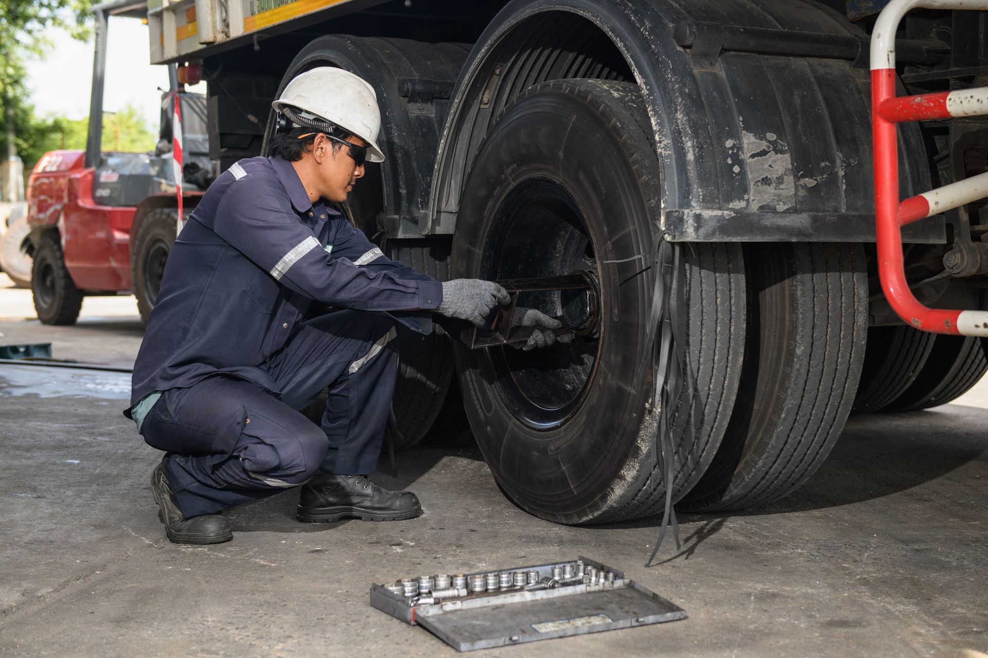 Mechanic in blue uniform inspects truck tires, squatting near wheels; a red forklift is in the background.