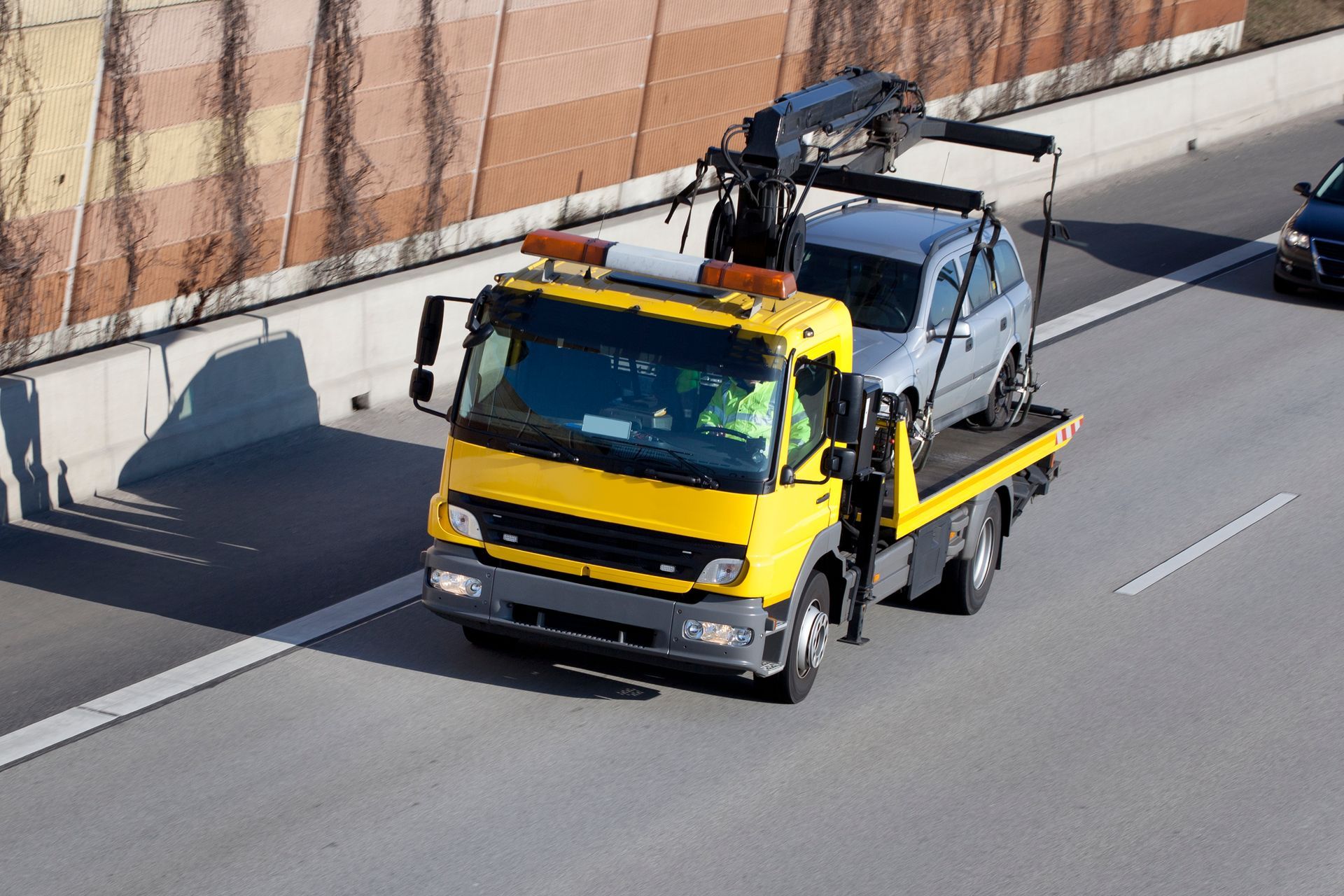 Yellow tow truck carrying a gray car on a highway, next to a retaining wall.