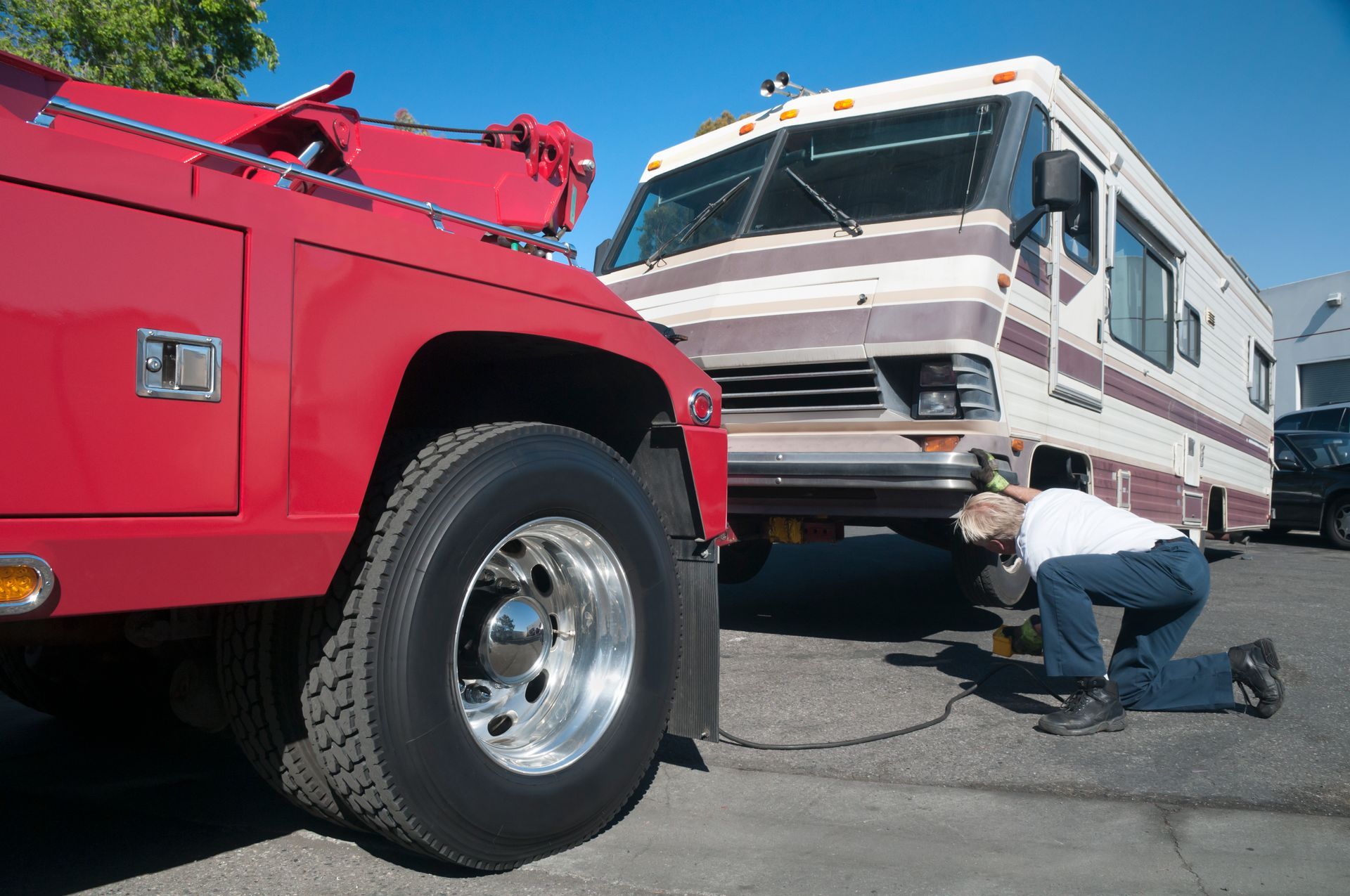 Red tow truck towing a beige and brown RV; a person kneels, working on the RV tire.