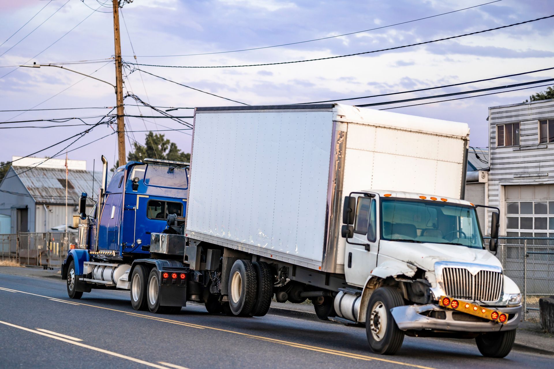 Blue semi-truck towing a white box truck, damaged front end, parked on a road, rural setting. Blue semi-truck towing a white box truck, damaged front end, parked on a road, rural setting.