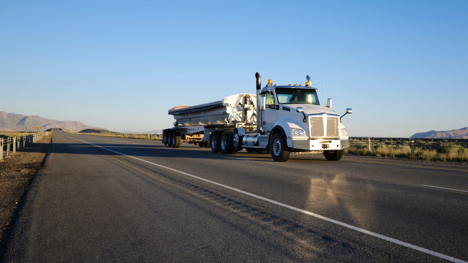 White semi-truck driving down a paved road, mountains in background under a clear blue sky.