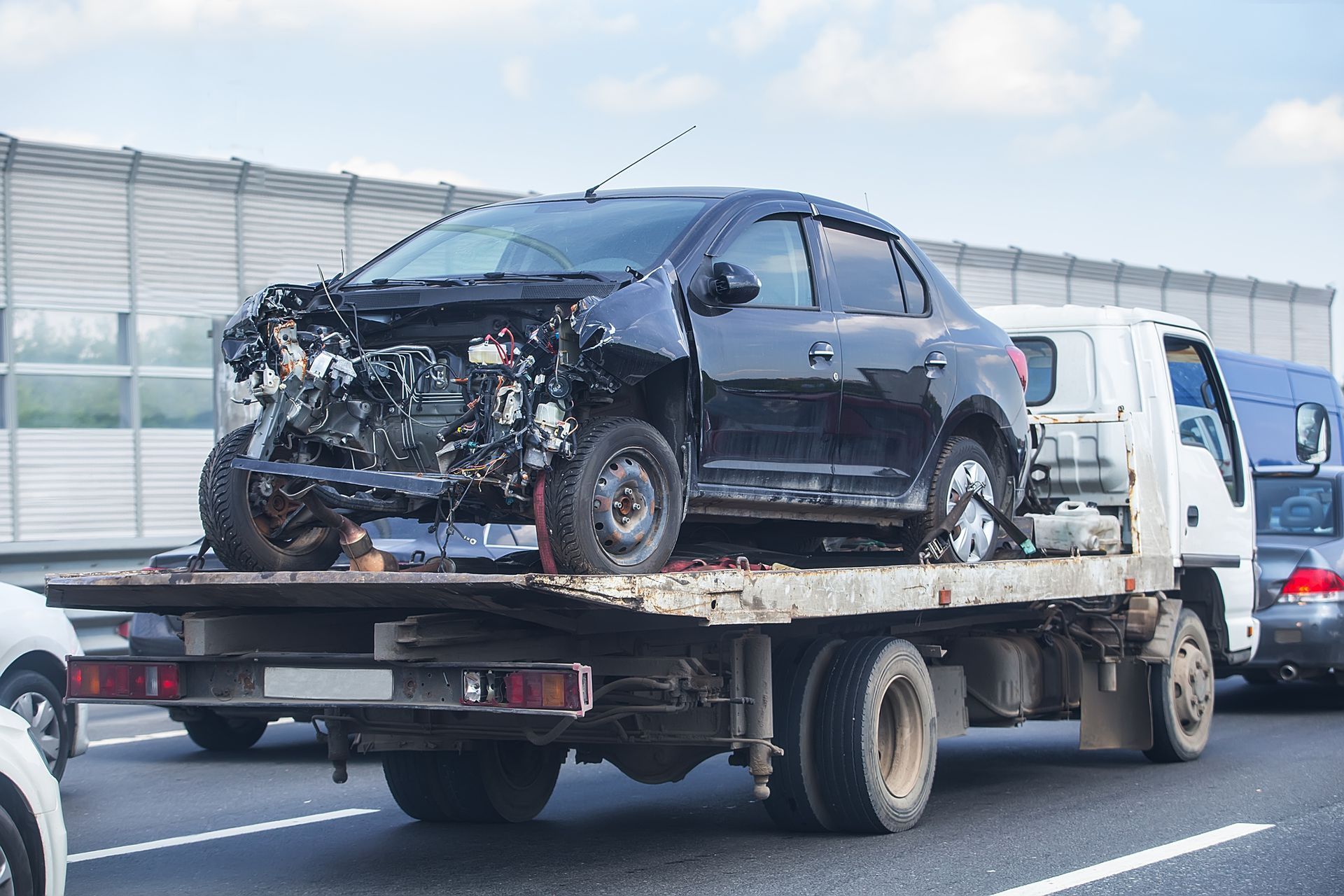 A severely damaged black car being towed on a flatbed truck on a highway.