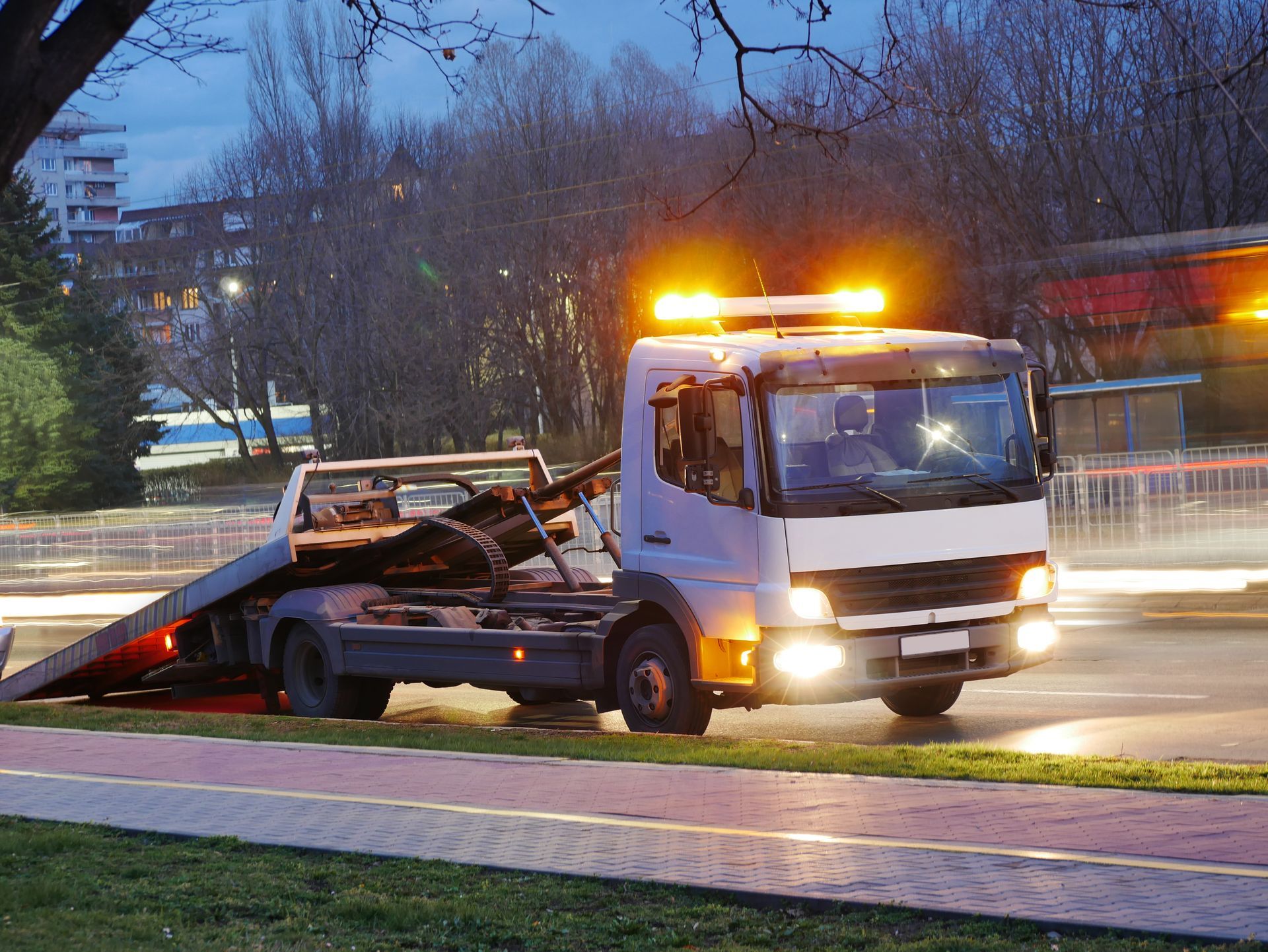 Tow truck with flashing amber lights on the side of a road at dusk.