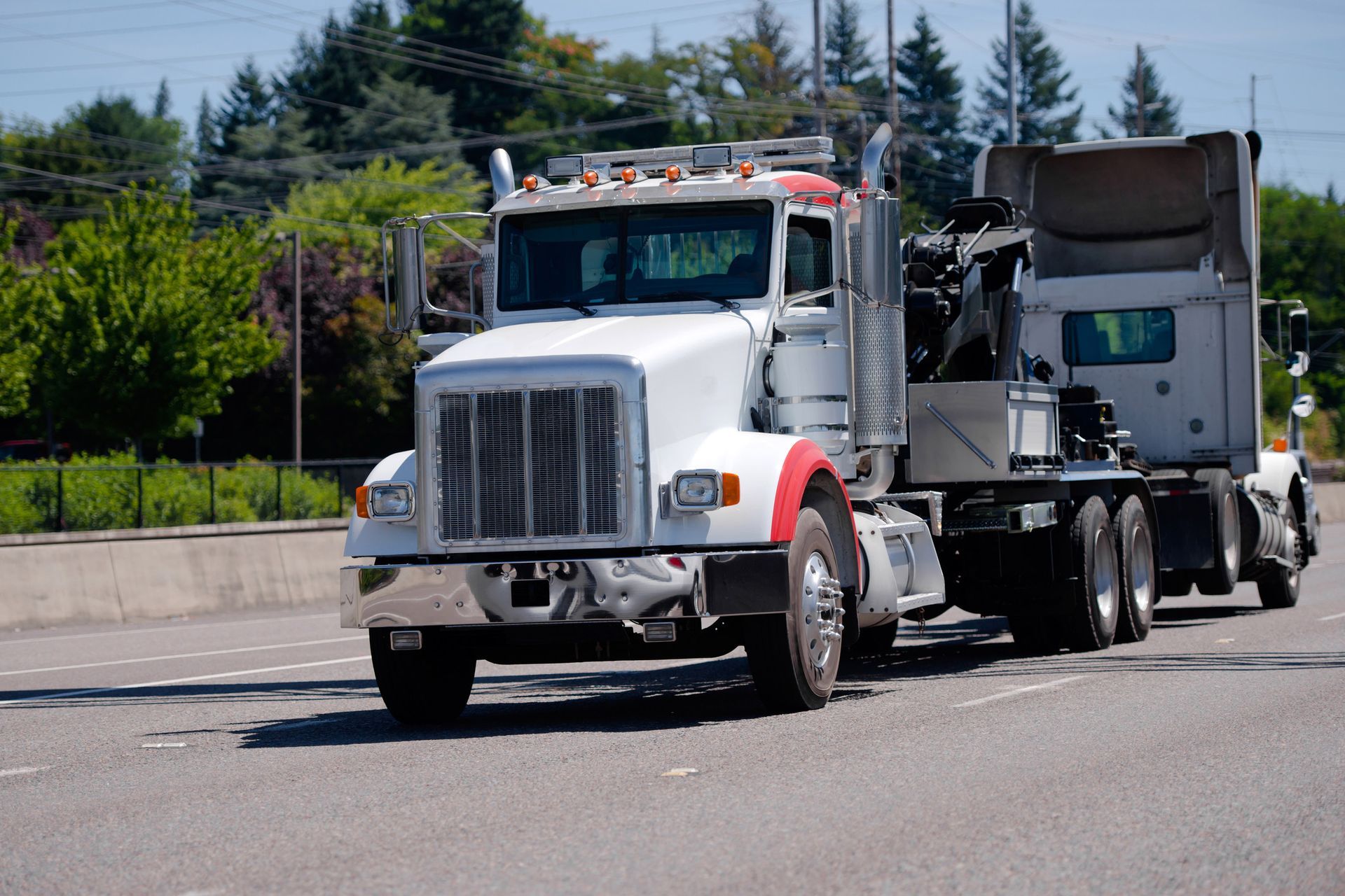 White semi-truck driving on a paved road, red accents on the cab, trees in the background.
