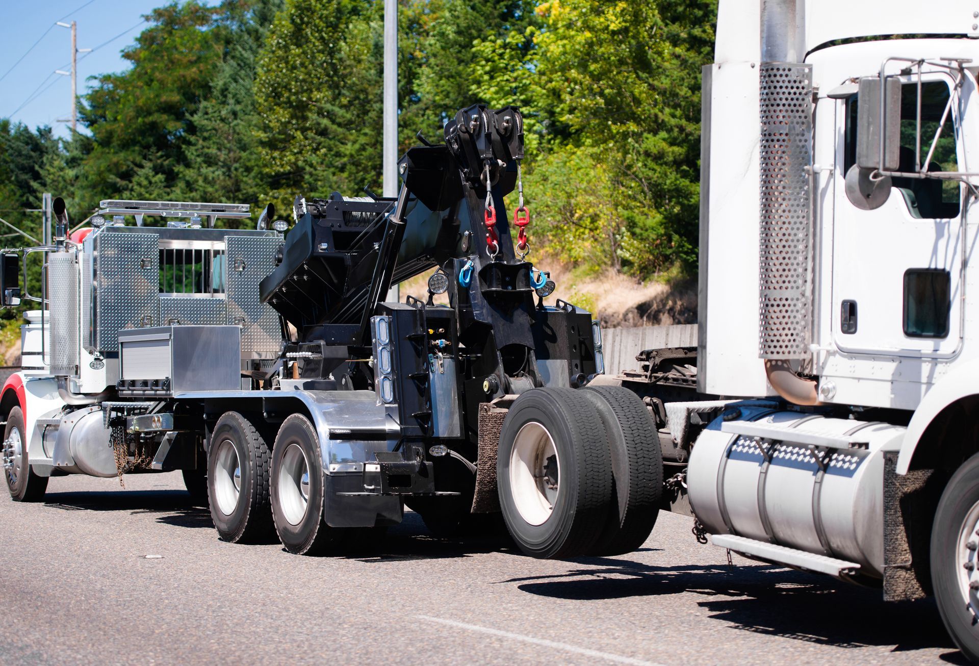 Tow truck towing a semi-truck on a road. The tow truck is black and white, and the semi is white. Sunny day.