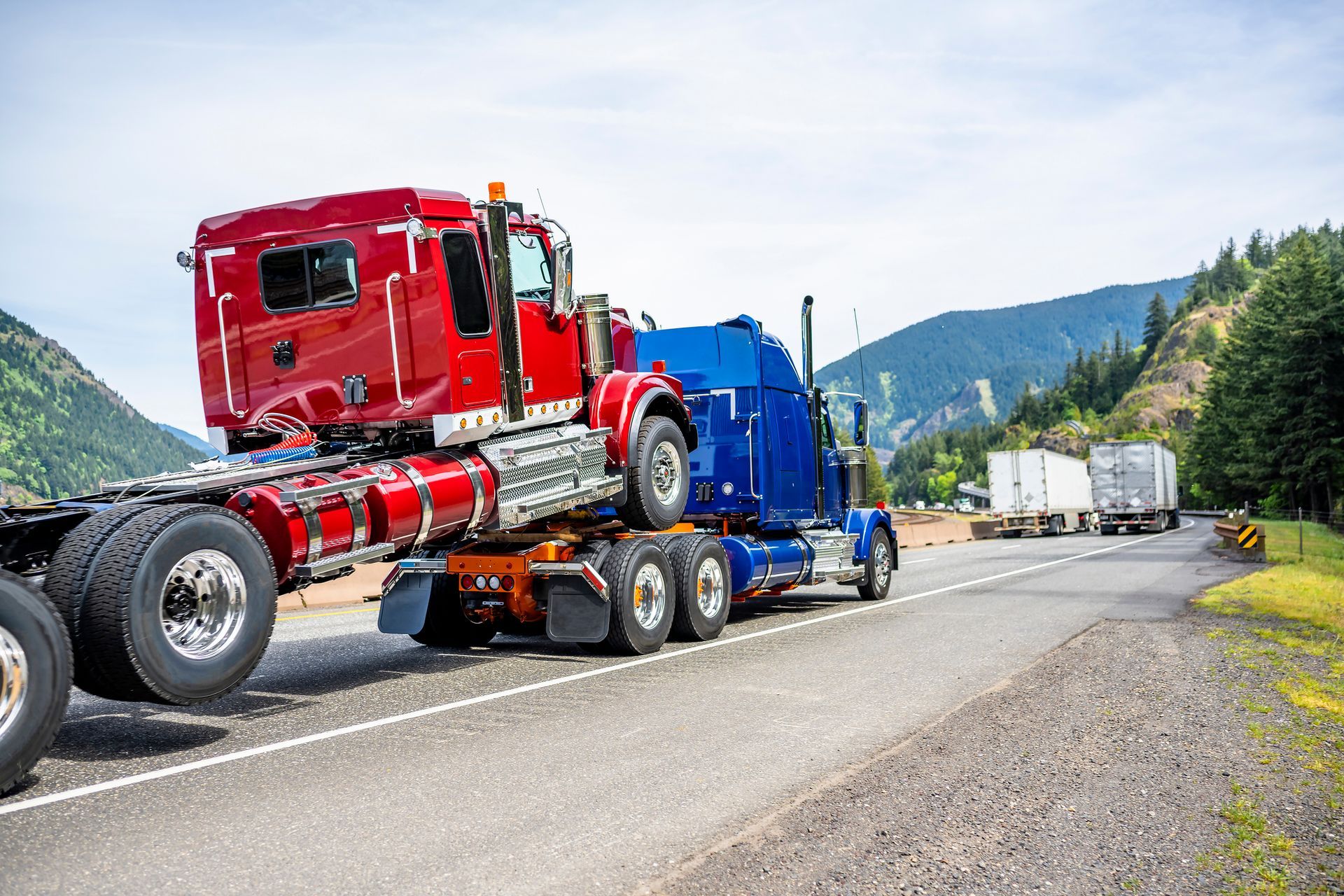A powerful blue big rig semi-truck transporting a red semi-truck tractor on the highway.