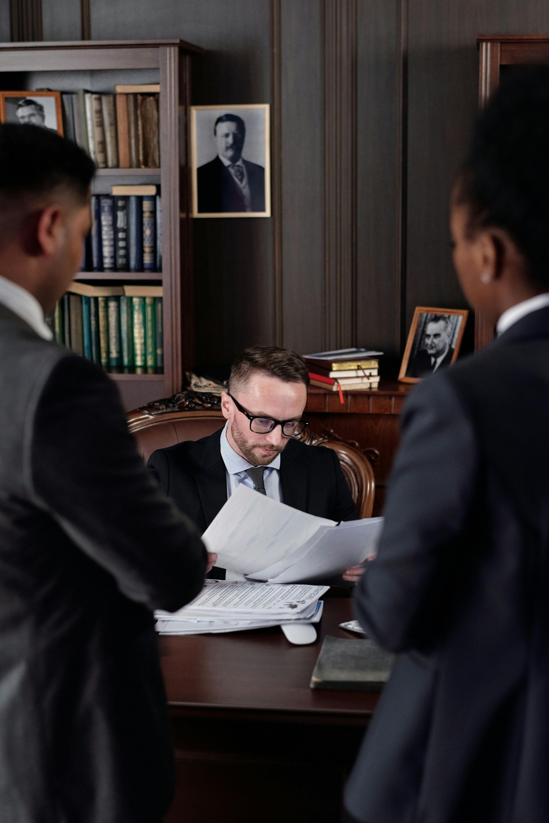 Three people in suits in an office. A man sits at a desk, reviewing papers. Two others stand nearby.