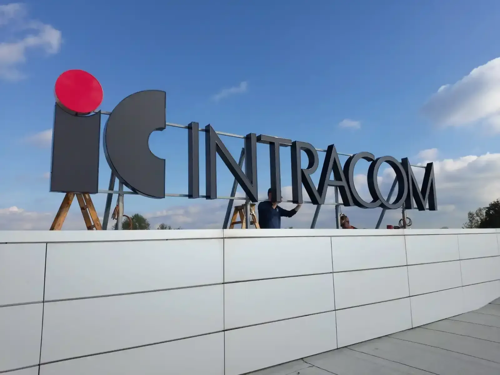 Insegna INTRACOM su un tetto contro un cielo azzurro, con logo circolare rosso e parete bianca di fronte.
