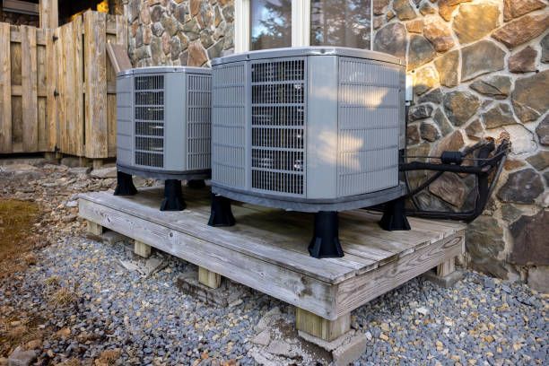 Two outdoor HVAC units on a wooden platform beside a stone wall and fence