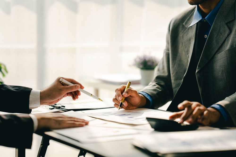 Two business professionals collaborate at a desk, reviewing documents and using a calculator in a bright office.