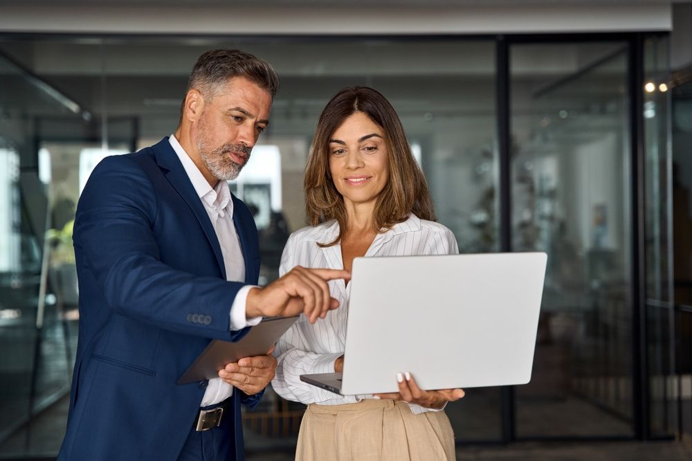 Mature Latin business man and European business woman discussing project on laptop gadget in office. Two diverse partners, colleagues, team of confident professional business people work together.