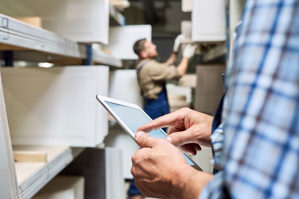 Person using a tablet in a warehouse, another person in the background reaching for boxes on shelves.