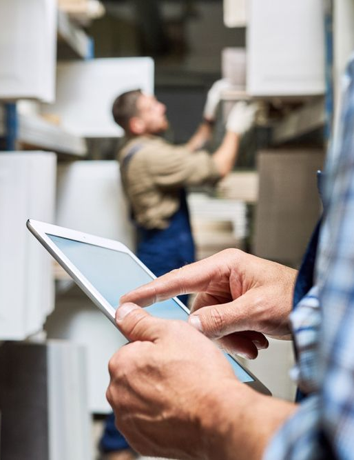 Man using a tablet in a warehouse, another man in the background arranging boxes on a shelf.