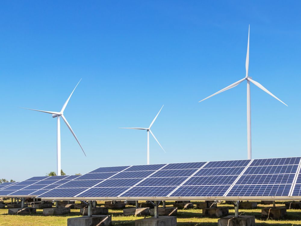 A field of blue solar panels set against a clear blue sky with three white wind turbines in the background.