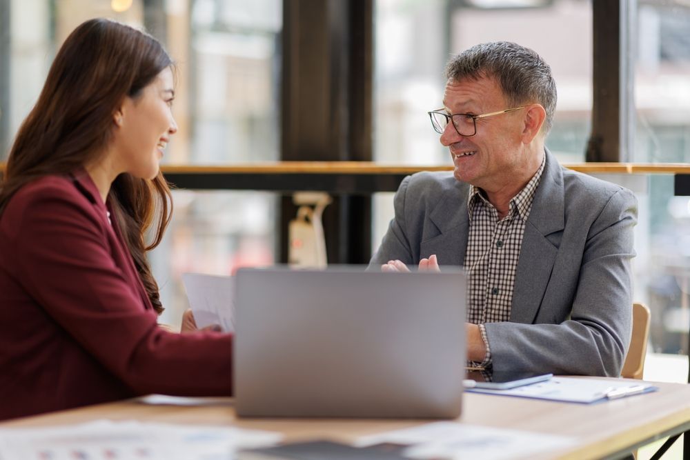 Woman and man in business attire at a table with laptop, documents, and smiling.