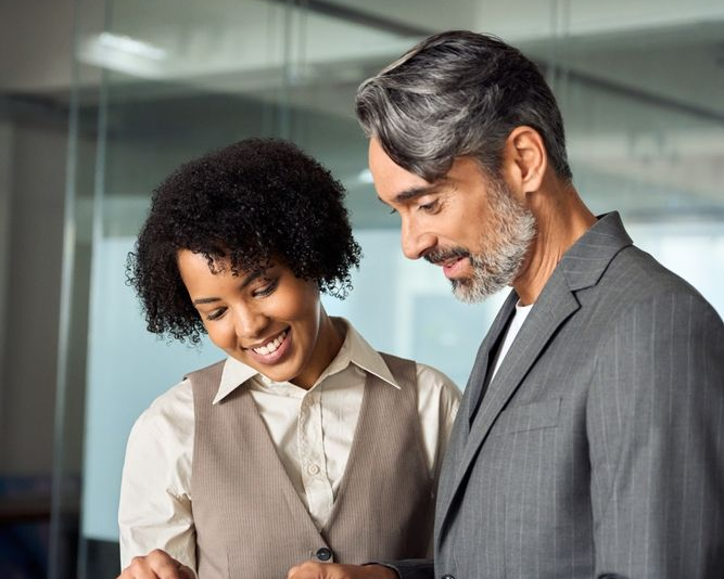 Woman and man looking at a tablet, both smiling. They are in a modern office with glass walls.