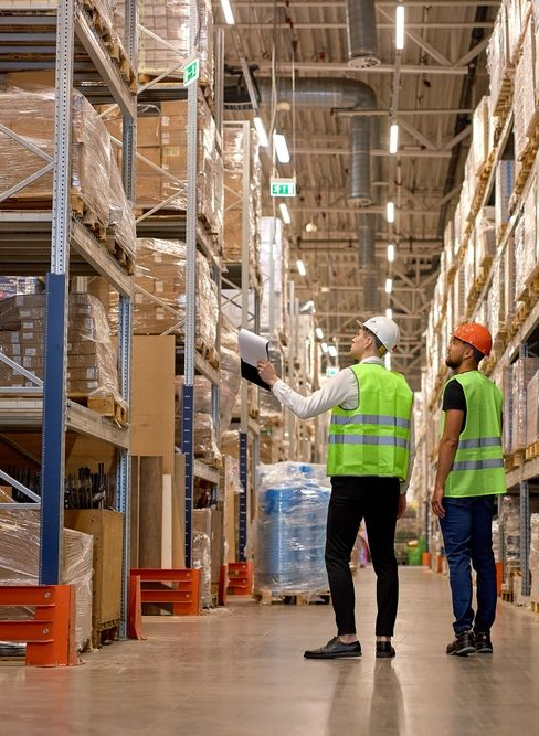 Two warehouse workers in safety vests and hard hats inspect tall pallet racks in an aisle.