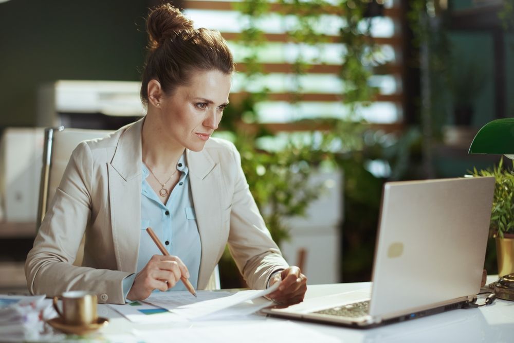 Woman in blazer at a desk, looking at paperwork and laptop, with plants in the background.