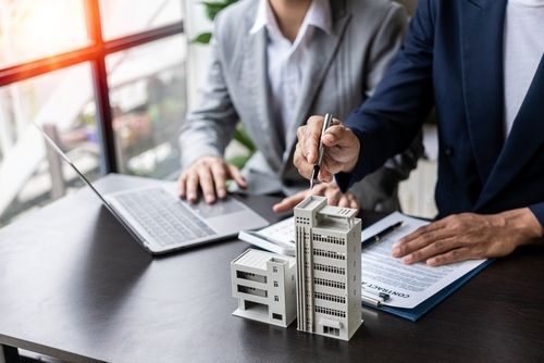 Two people in suits pointing at miniature buildings on a table, with a laptop and documents.