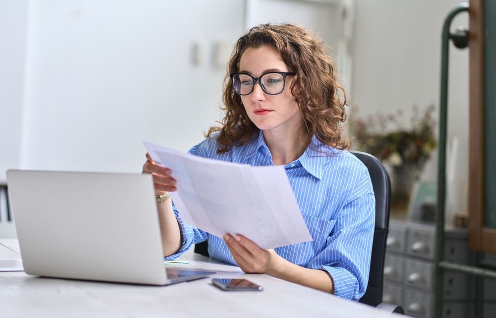 A woman is sitting at a desk looking at a piece of paper in front of a laptop computer.