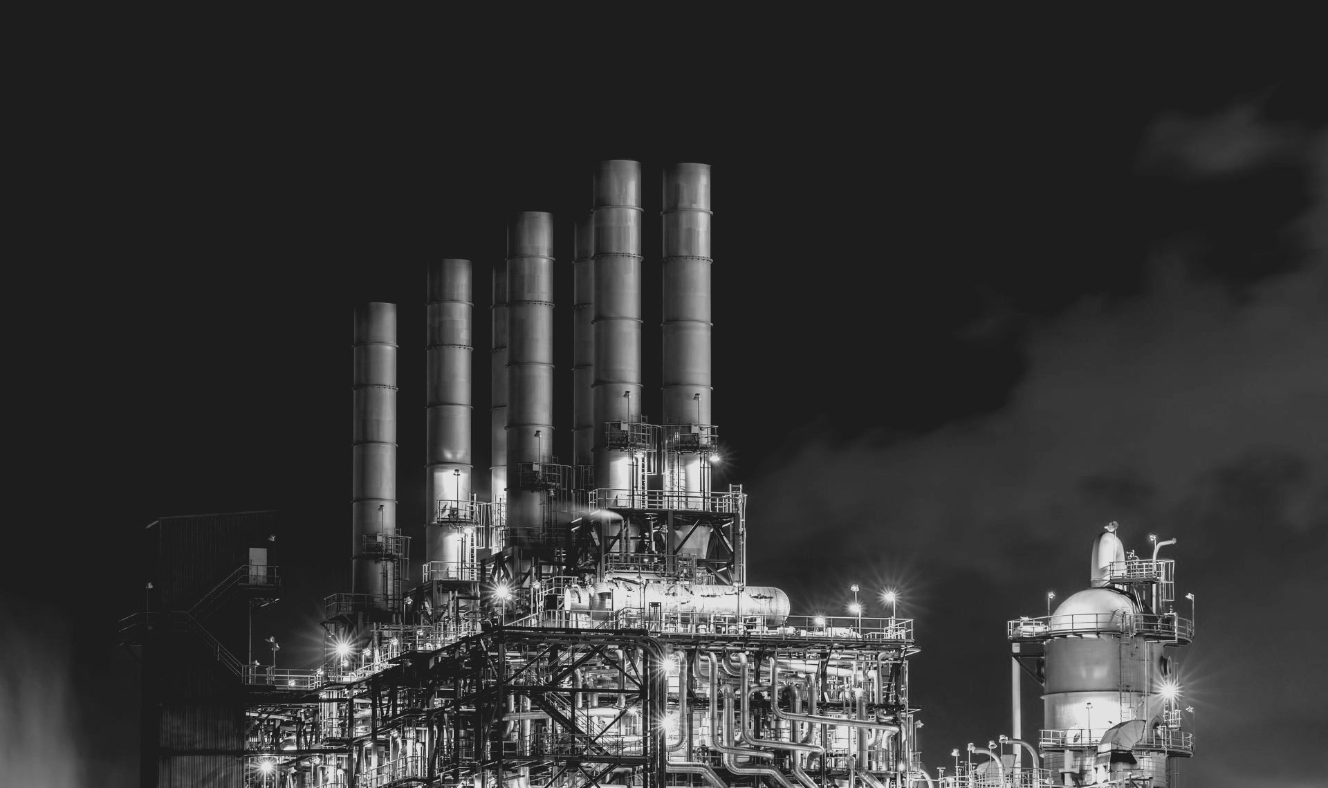 Black and white nighttime photograph of an industrial oil refinery with multiple smokestacks, emitting smoke.