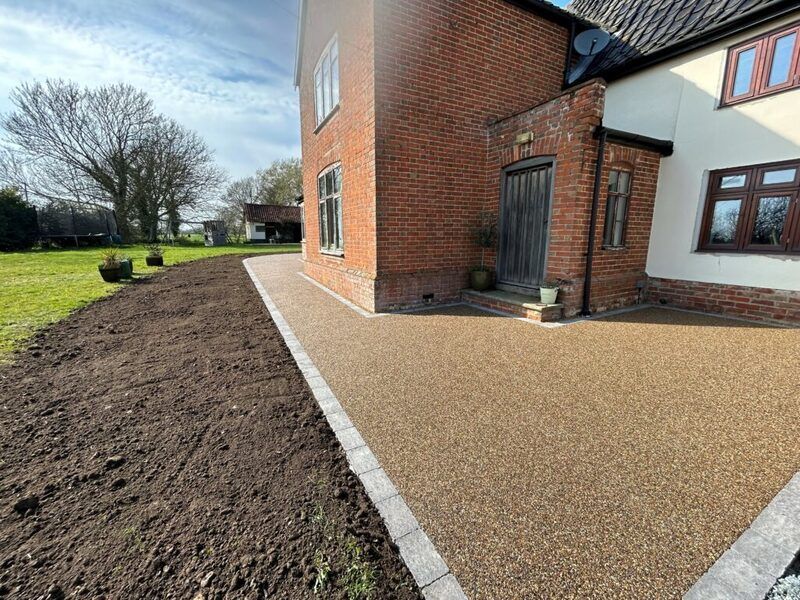 A brick house exterior with a newly laid, light gray tiled patio leading to a dark fence and a child's toy in the yard.