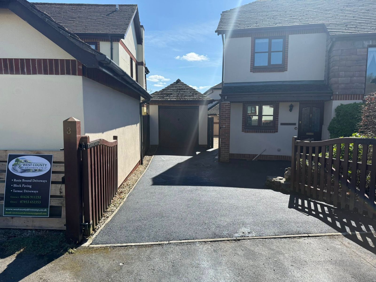 A paved driveway leads toward a garage situated between two light-colored, two-story houses under a clear blue sky.