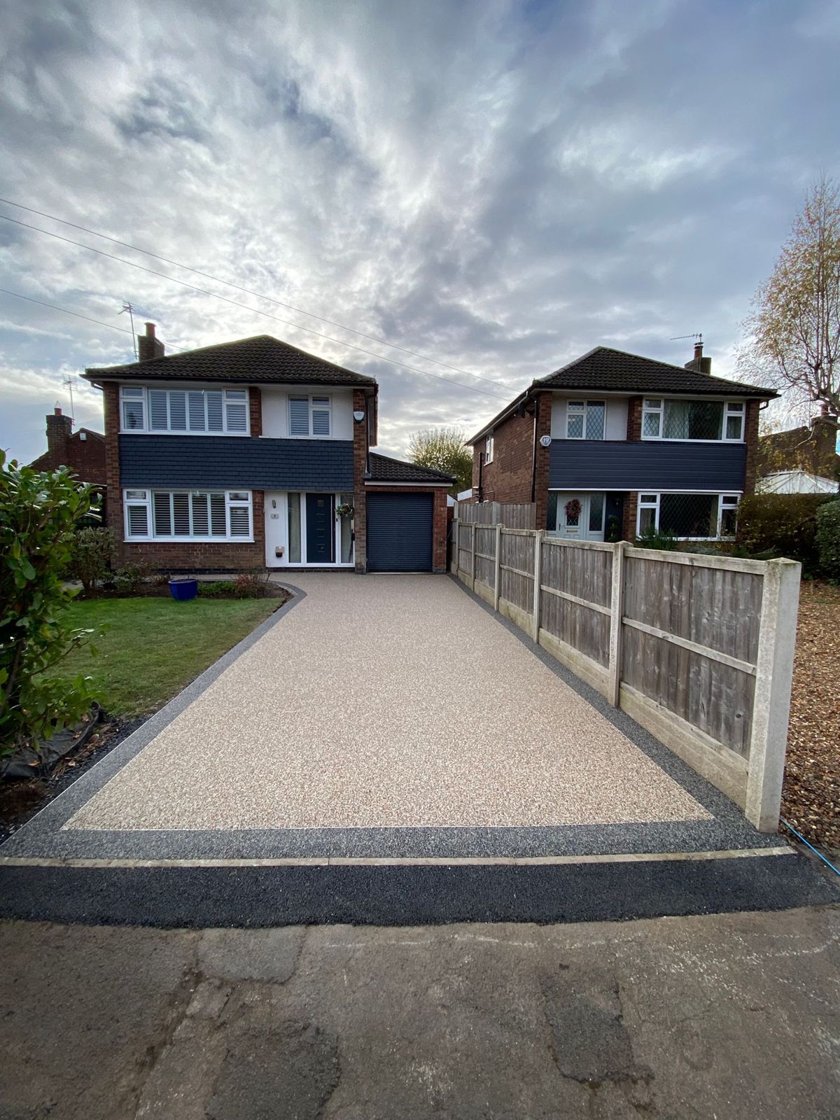 A newly paved driveway with beige resin and dark borders leading to two detached brick houses under a cloudy sky.
