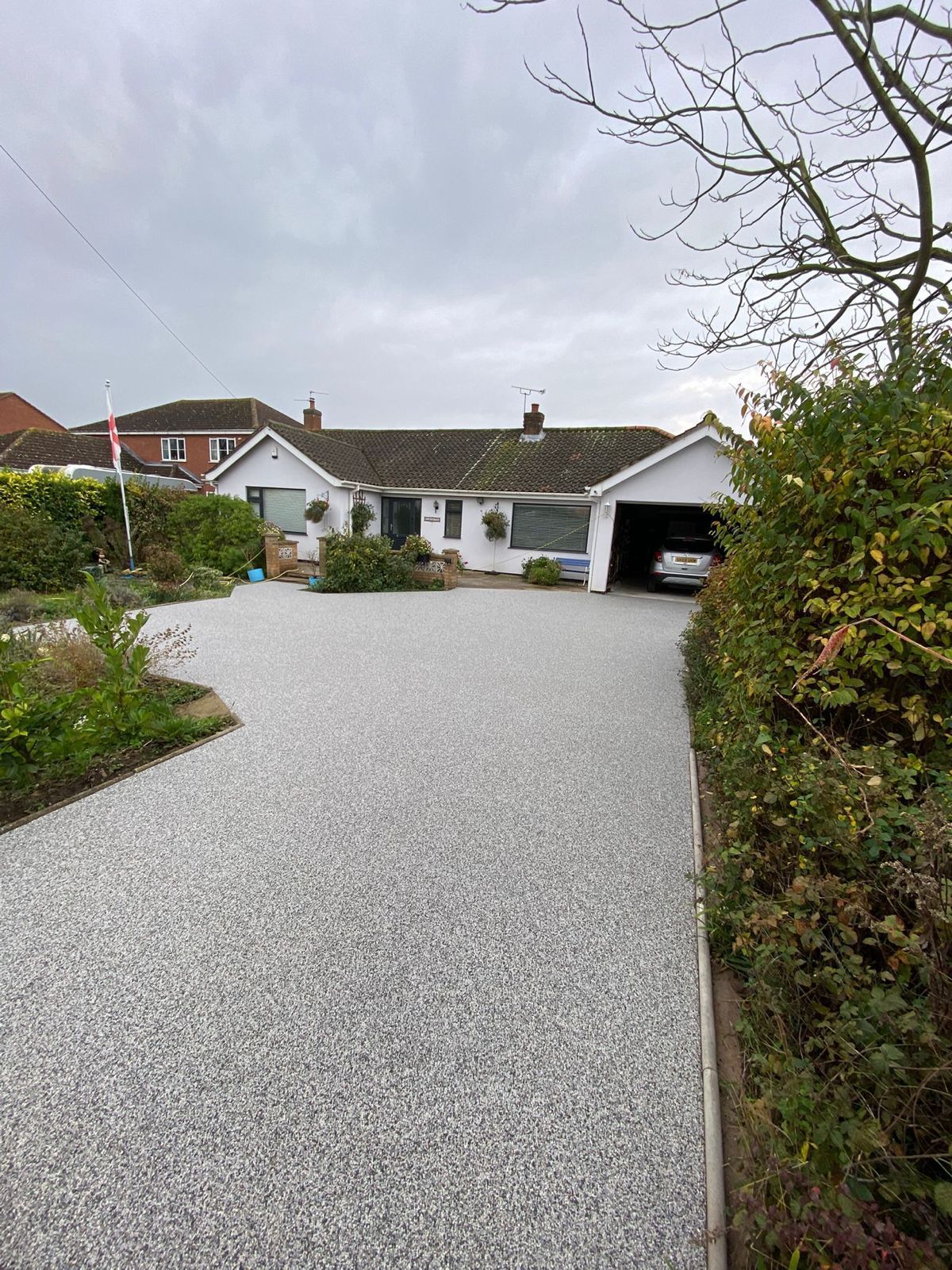 A white, single-story house with a grey tiled roof and a large gravel driveway, framed by bushes under a cloudy sky.