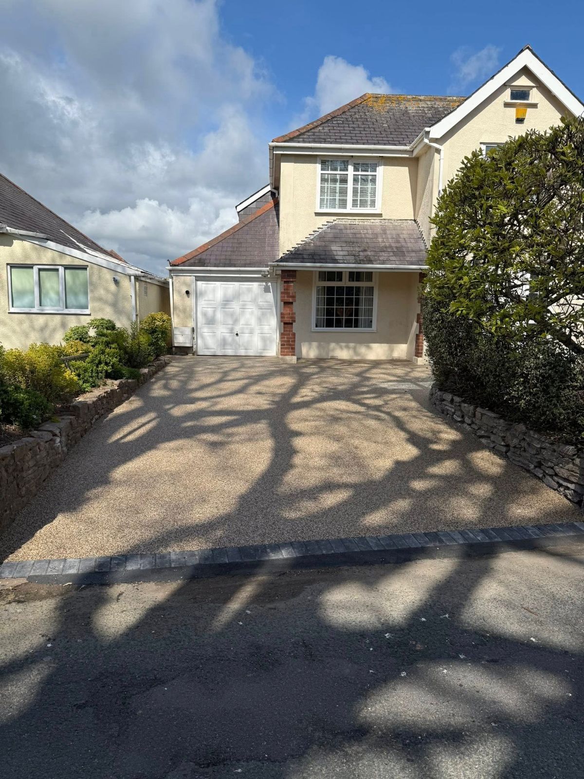 A beige two-story house with a white garage door and a gravel driveway, framed by shadows and greenery under a blue sky.