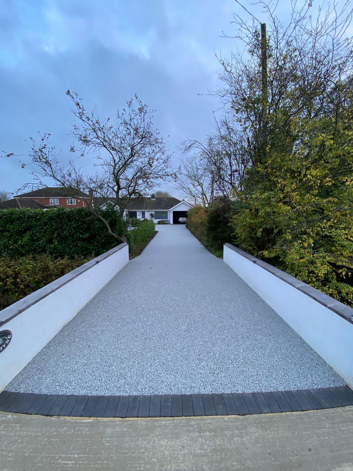 A two-story suburban house with a beige exterior, a white garage door, and a gravel driveway under a bright sky.