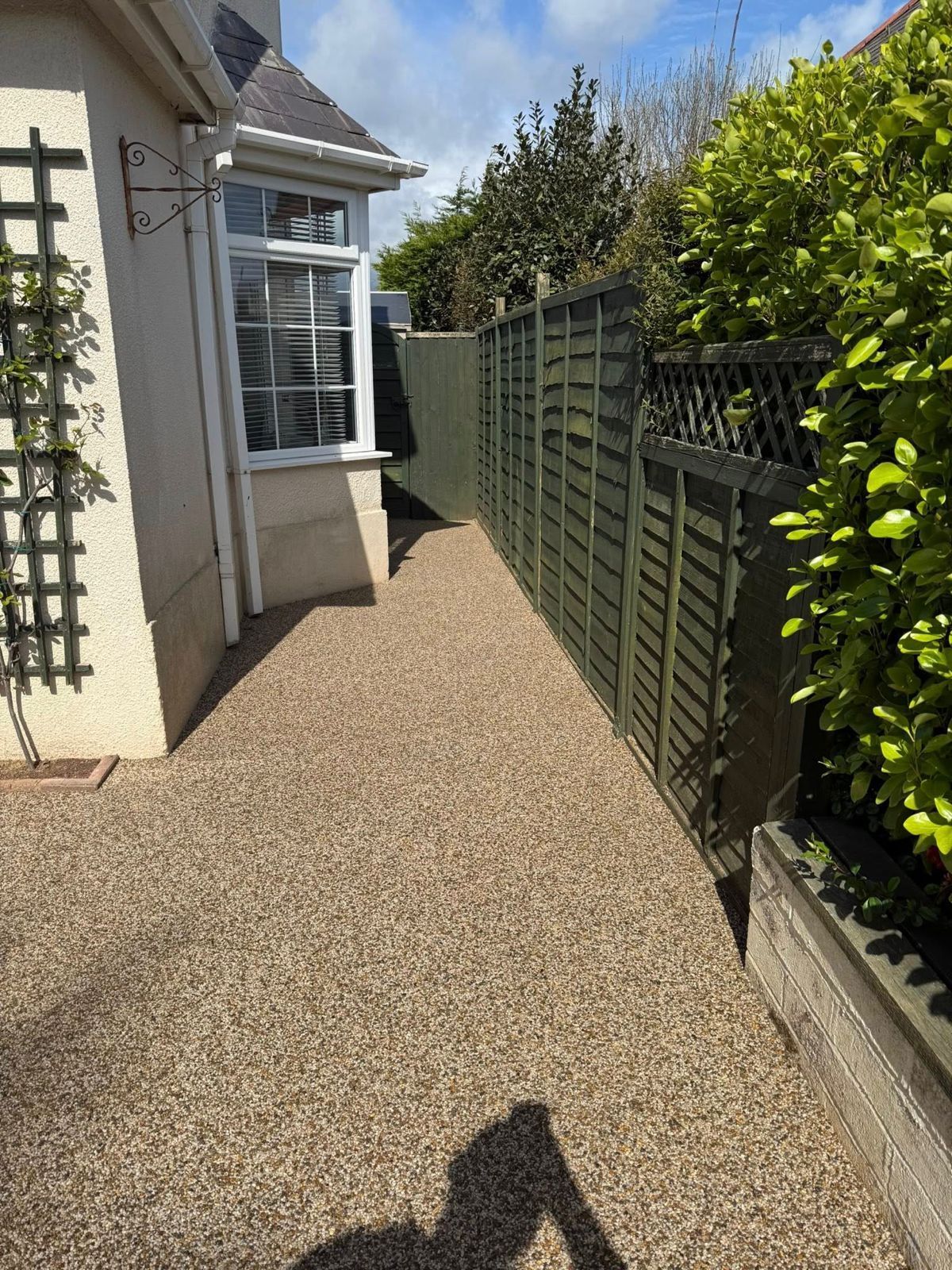 A narrow pathway with a resin-bonded gravel surface, running beside a white house wall and a dark wooden garden fence.