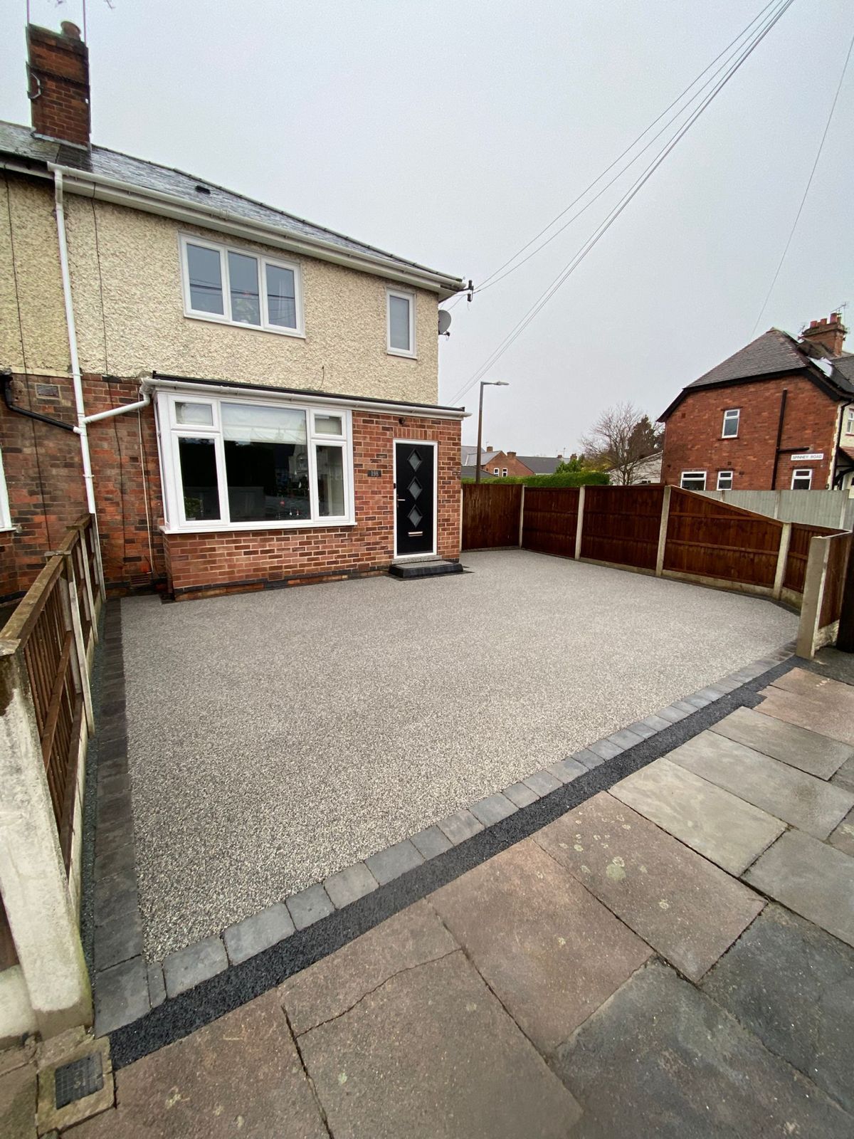 A beige house with a glass-paneled door and bay window faces a gravel driveway next to a grassy lawn and a green hedge.