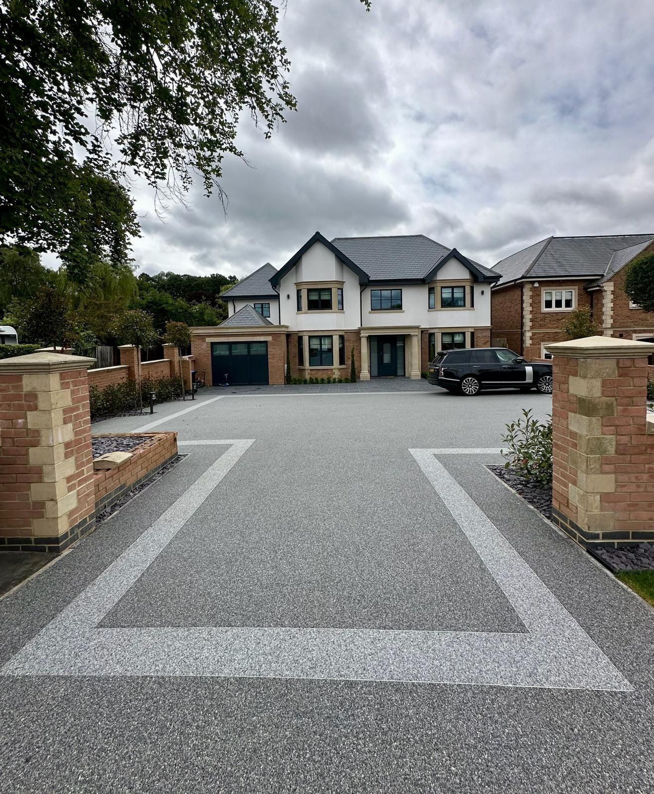 A modern grey resin driveway with a decorative light-colored paved border in front of a brick house with a dark door.