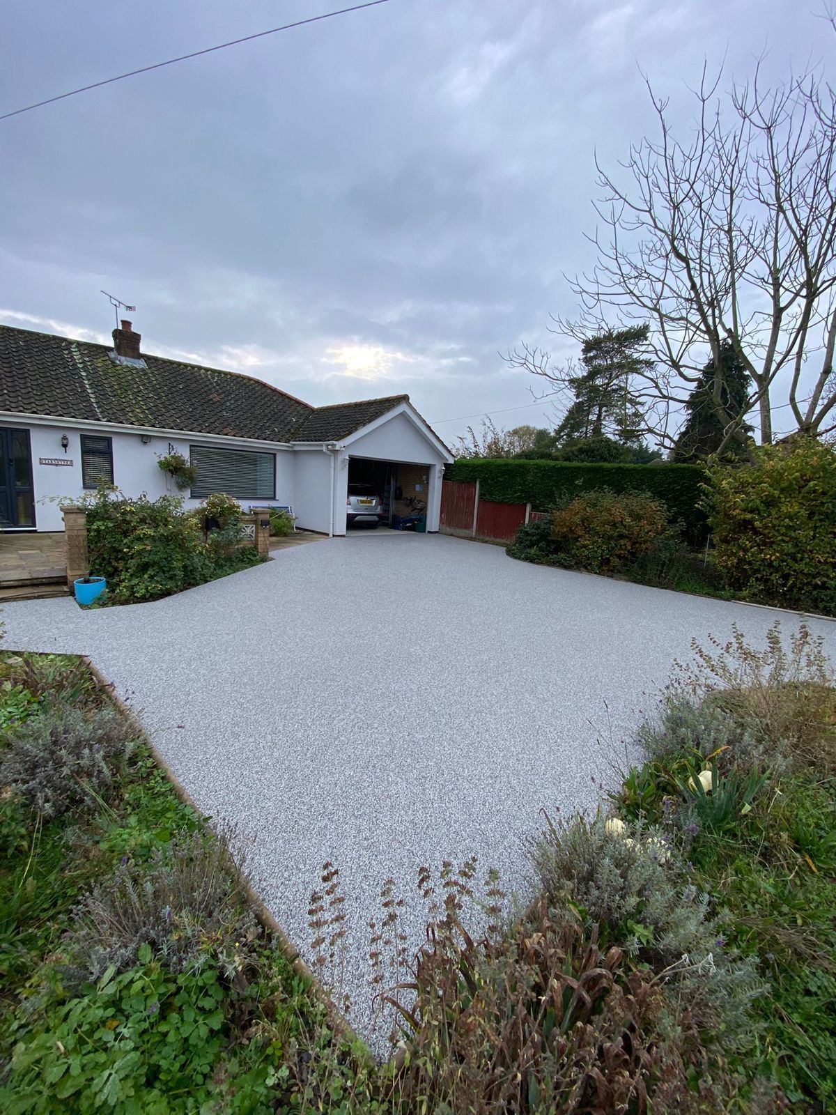 A light gray gravel driveway leads to the garage of a white single-story house under a cloudy sky.