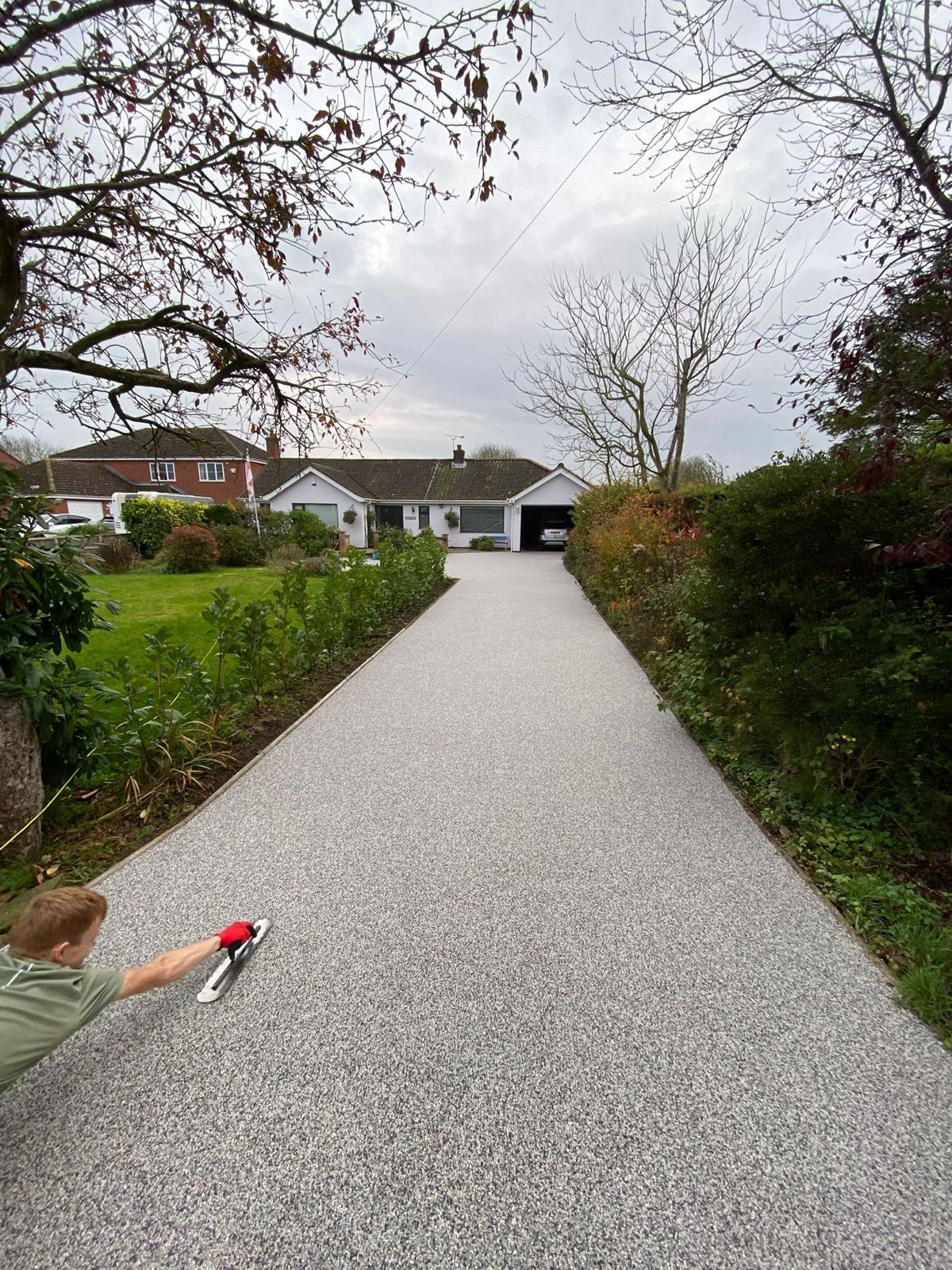 A person in a light green shirt applies resin to a long, gray pebble driveway leading to a house.