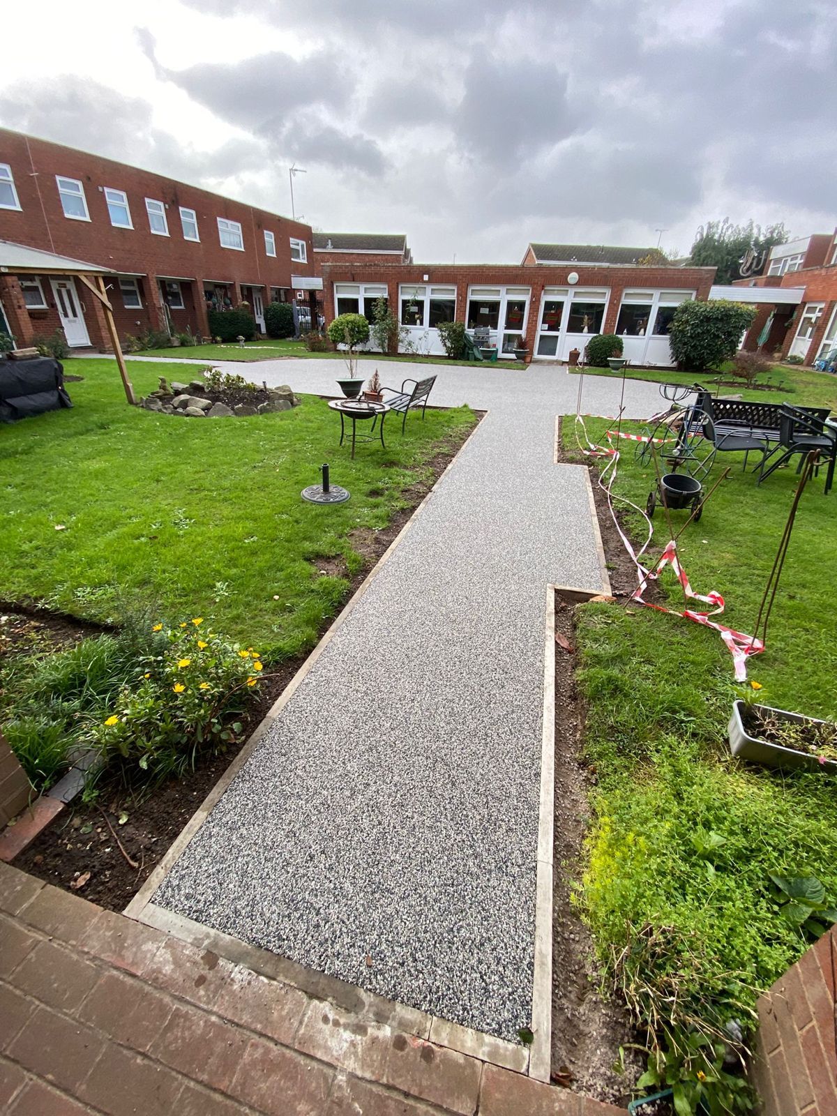 A new gravel path leads through a green courtyard between two low-rise red brick buildings under a cloudy sky.