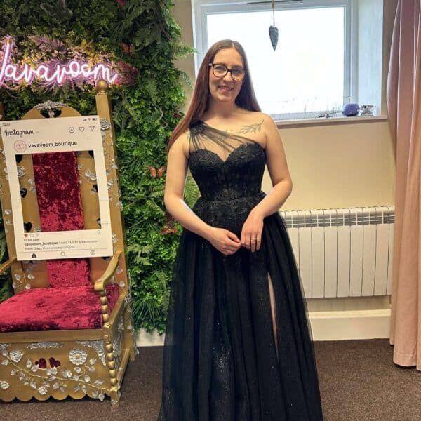 Woman in black formal gown stands near throne and floral wall.